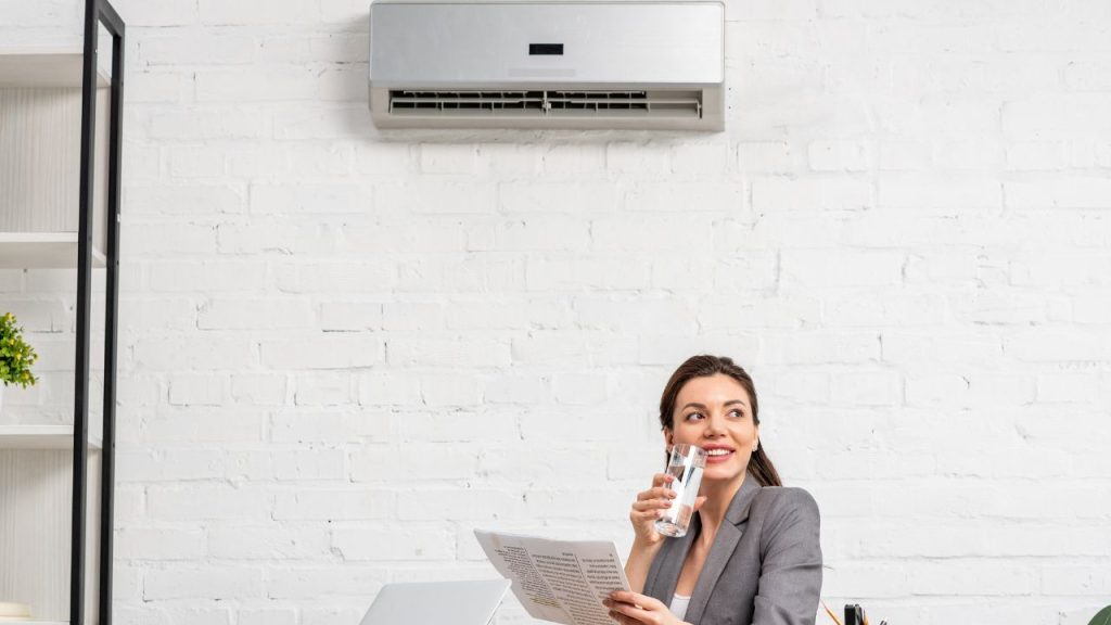 a happy woman sitting on a couch and drinking water with a new air conditioning unit behind her